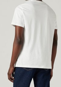White cotton t-shirt with short sleeves and a round neck. Features a small branded tag at the hem. Worn with dark blue jeans.