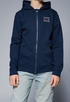 Osoba ubrana w granatową bluzę rozpinaną Tommy Hilfiger z założonym kapturem oraz jasnoniebieskie dżinsy, stojąca na tle jednolitego tła.