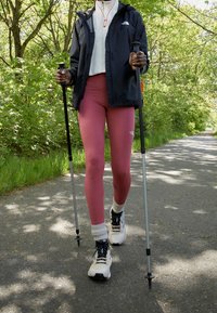 Nordic walking gear: person in a black jacket and white top, pink leggings, white socks, and sports shoes, using trekking poles on a path.