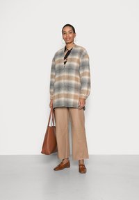 Cardigã oversized xadrez em bege, cinza e branco, com decote em V e ombros caídos, combinado com calças largas em tom de areia e sapatos castanhos.