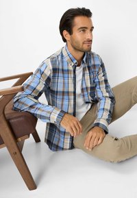 Gestrikte shirt in blauw, bruin en beige, met een button-down kraag en lange mouwen. Draagt over een witte T-shirt, gecombineerd met beige broek.
