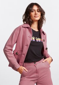 Chaqueta de algodón rosa con cuello y botones en la parte frontal, combinada con una camiseta negra con texto en arcoíris. La modelo posa con una mano en el bolsillo.