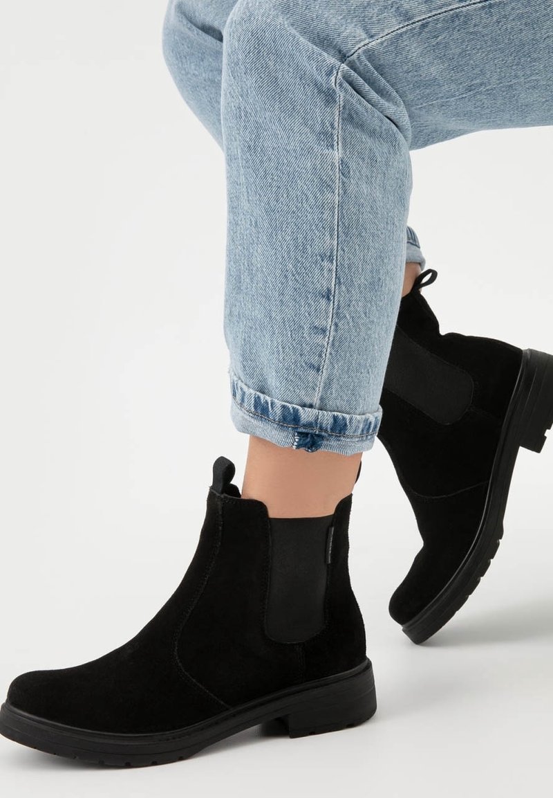 Personne portant un jean bleu clair retroussé et des bottines en daim noir avec des panneaux élastiques sur les côtés, debout sur une surface blanche.