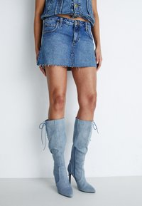 Jeansminikjol med trasig fåll och en figursydd design, kombinerad med ljusblå knähöga stilettkängor med snörningsdetaljer.