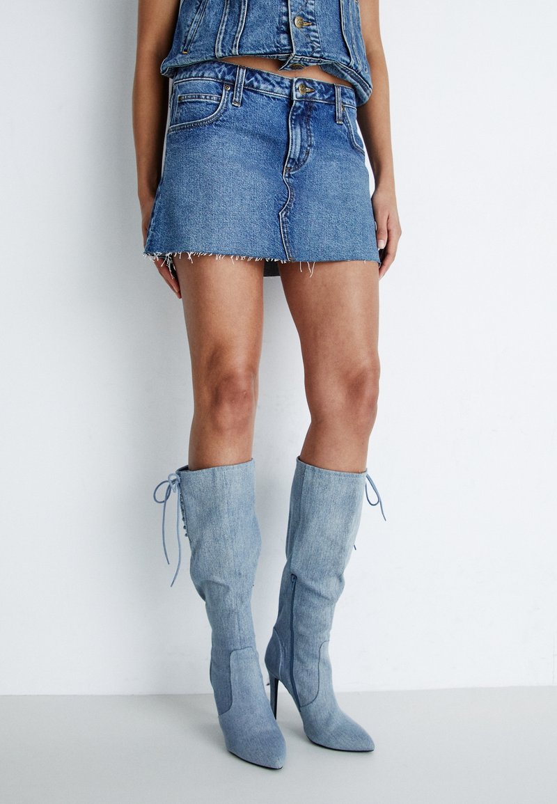 Jeansminikjol med trasig fåll och en figursydd design, kombinerad med ljusblå knähöga stilettkängor med snörningsdetaljer.