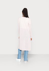 Long pink cardigan with long sleeves, made of soft fabric. Worn over light blue jeans and white shoes, with a relaxed fit and simple design.