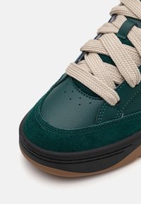 Zöld bőr és velúrból készült sneaker bézs fűzővel, kerek orral �és fekete talppal. Az orr részén perforációk találhatók, valamint kontrasztos varrás.