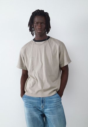 Joven con piel oscura y rastas, lleva una camisa beige de manga corta y vaqueros azul claro, de pie con las manos en los bolsillos.