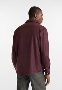 Bordeaux kleurige knoopshirt met kraag, lange mouwen en details van een borstzak. Gemaakt van een textuurstof, voorzien van goudkleurige knopen.