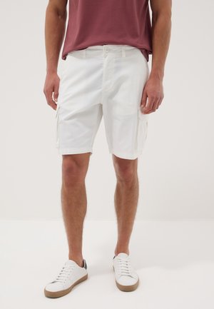 Mann trägt weiße Cargo-Shorts und weiße Turnschuhe, steht vor einem schlichten hellen Hintergrund.