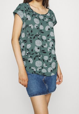 Vrouw die een groen topje met korte mouwen met witte bloemenprints en een blauwe denim minirok draagt tegen een effen lichte achtergrond.