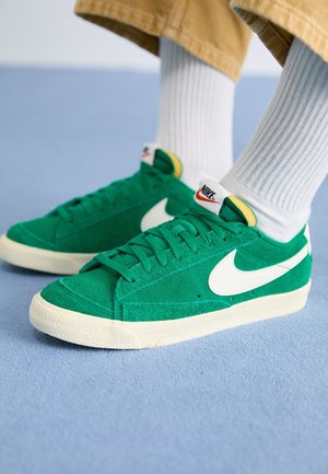 Persona con calcetines blancos acanalados y zapatillas verdes de ante Nike con el logo blanco y suelas crema sobre una alfombra azul.