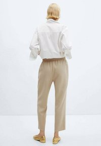 Weiße Baumwollbluse mit hochgekrempelten Ärmeln, beigen elastischen Hosen und goldenen Slippern, von hinten gesehen vor einem einfarbigen Hintergrund.