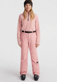 Γυναίκα με ροζ σαλοπέτες, αντίστοιχο ροζ μακρυμάνικο μπλουζάκι, μαύρη ζώνη, μαύρες μπότες σκι και μαύρη κορδέλα στο κεφάλι, όρθια και κοιτάζοντας μπροστά.