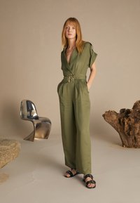 Olivově zelený overal s překříženým designem, krátkými rukávy a širokým opaskem s kulatou sponou. Stylizováno s černými sandály, umístěno na neutrálním pozadí.