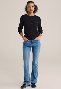 Crni teksturirani kardigan s okruglim izrezom, zatvaranjem na gumbe i rebrastim manšetama, uparen s plavim trapez trapericama i crnim tenisicama.