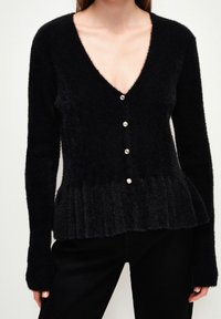 Czarny kardigan z puszystego materiału, z głębokim dekoltem w kształcie litery V, pięcioma guzikami perlami i peplum z teksturą prążkowaną. Długie rękawy.