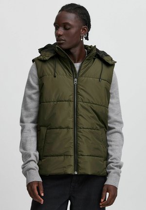Olivgrüne gepolsterte Weste mit Reißverschluss, abnehmbarer Kapuze und Seitentaschen, über einem hellgrauen Pullover mit gerippten Bündchen getragen.
