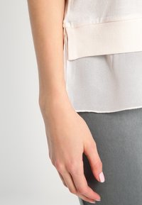 Jasno beżowa bluzka z teksturowanej tkaniny z miękkim, przezroczystym dołem. Szare spodnie o gładkim wykończeniu, z częściowo widoczną ręką spoczywającą na nich.