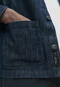 Nærbillede af en hånd, der hviler i en lomme på en mørk, stribet denimjakke med synlige knapper og stoftekstur.