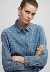 Camicia in denim con tessuto blu chiaro, caratterizzata da accenti blu sparsi. Presenta un colletto classico e polsini con bottoni.