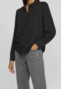 Černá košile s límečkem, dlouhými rukávy a jemnou texturou. Nosená s šedými džíny s vysokým pasem, vytvářející neformální outfit.