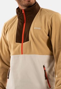 Sudadera de forro polar en beige y blanco con el pecho marrón, presenta una cremallera naranja, dos bolsillos laterales y un logo en el pecho.