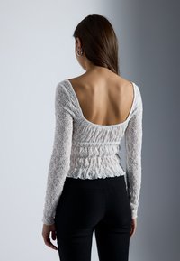 Paita dantelă cu mâneci lungi și ajustate, spate deschis larg și textură plisată. Culoare gri deschis cu modele intricate, asociată cu pantaloni negri.