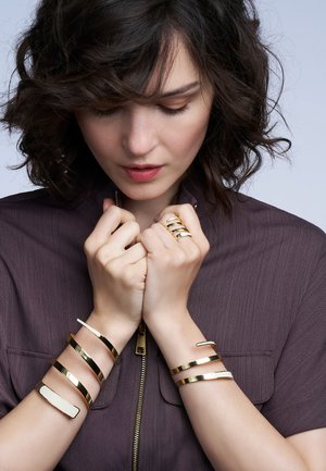 Femme aux cheveux bouclés et foncés, portant un haut zippé marron et plusieurs bracelets et bagues dorés et couleur crème, regardant vers le bas avec les mains près de son visage.