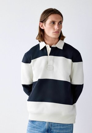 Junger Mann mit schulterlangem braunem Haar trägt ein schwarz-weiß gestreiftes Rugbyshirt und blaue Jeans, schaut nach rechts vor einem schlichten Hintergrund.