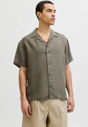Homme portant une chemise olive ample à manches courtes boutonnée et un pantalon beige, debout devant un fond clair uni, regardant vers la droite.