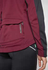 Bordeaux stoffen jack met zwarte accenten, voorzien van een ritszak en een contrasterend paneel. Glad oppervlak, sportieve pasvorm en logodetail aan de onderkant.