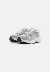 Scarpe da ginnastica grigie e bianche con tomaia in rete, dettagli in suede e suola in gomma texturizzata, dotate di lacci e dettagli riflettenti.