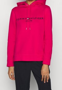 Różowa bluza z kapturem wykonana z miękkiego materiału, z kapturem ze ściągaczem, logo z przodu oraz ściągaczami na rękawach. W połączeniu z czarnymi spodniami.