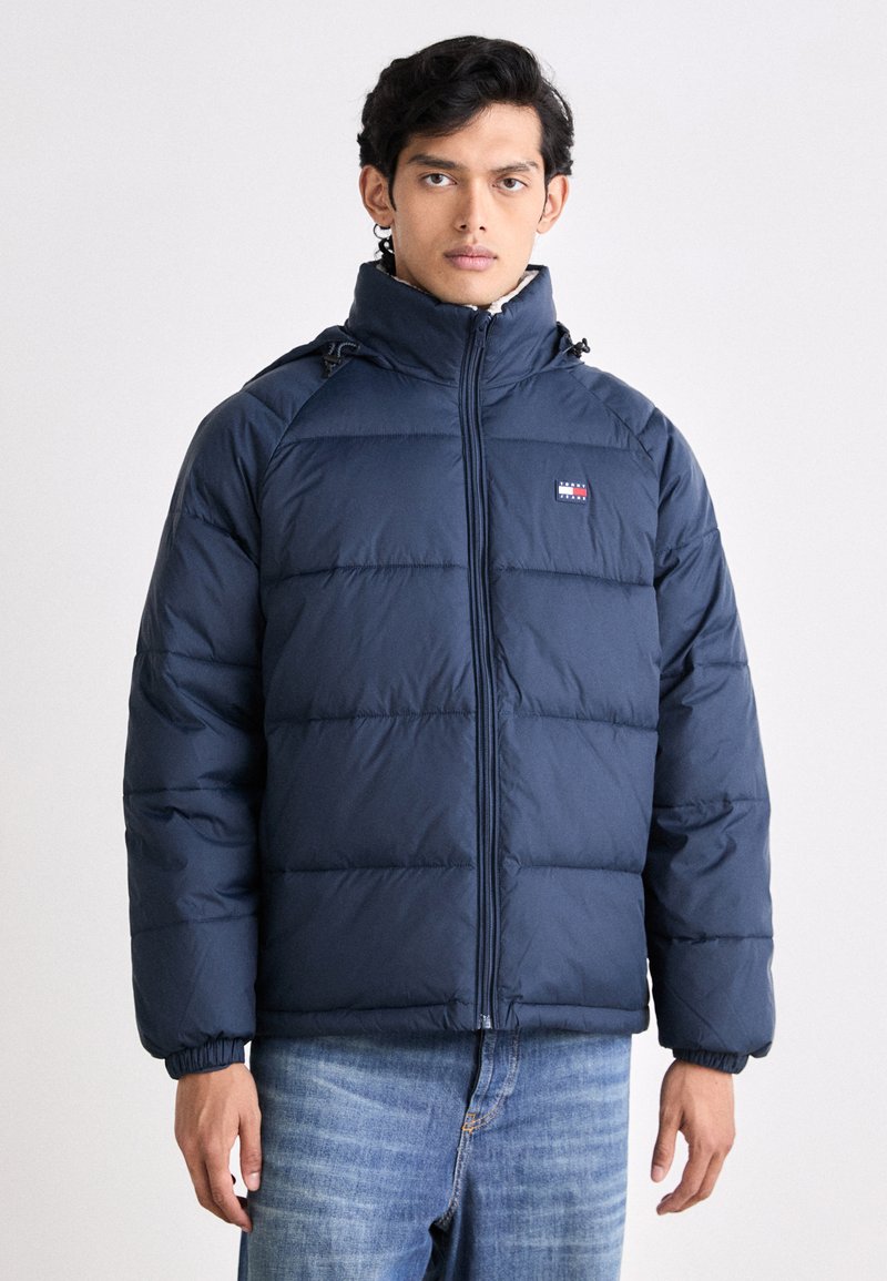 Chaqueta acolchada de color azul marino con cremallera en la parte delantera, puños elásticos y un cuello alto. Presenta acolchado horizontal y un pequeño logotipo de la marca en el pecho.
