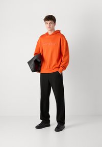 Felpa arancione con logo bianco, pantaloni neri sartoriali e sneakers nere. La felpa ha un cappuccio con cordino e polsini a coste.