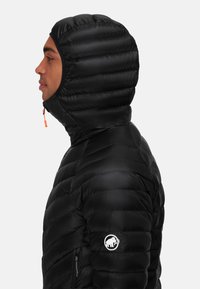 Zwarte pufferjack met een capuchon, voorzien van doorgestikte segmenten, een hoge kraag en een logopatch op de mouw. Ritsaccent in oranje.