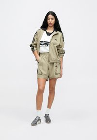 Béžová športová bunda s čiernymi pruhmi, zodpovedajúce šortky a biele tričko s čiernym textom. Strieborné športové topánky a biele ponožky.