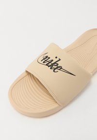 Sandale glissante beige avec un pied en relief et une large sangle arborant un logo Nike noir. Matériau lisse avec une forme arrondie.