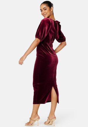 Bordeaux fluwelen midi-jurkje met pofmouwen, open rug met strikdetail, zijsplit en aansluitende silhoutte; gecombineerd met naakte heeled sandalen.