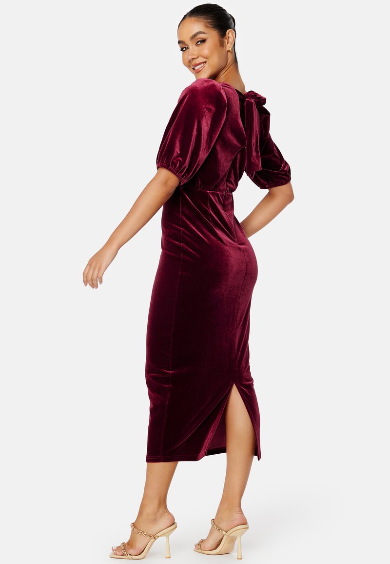 Abito midi in velluto burgundy con maniche a palloncino, dettaglio a schiena scoperta con fiocco, spacco laterale e silhouette aderente; abbinato a sandali con tacco nudi.