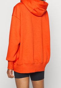 Överdimensionerad orange huvtröja med dragsko på huvan, ribbade ärmslut och nederkant, visad från baksidan, tillsammans med svarta shorts. Slät textur.