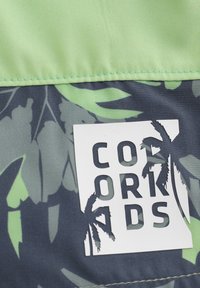 Färggamt tyg med gröna tropiska bladmönster mot en mörk bakgrund, med en vit märkesetikett som står "COORDS." Slät textur.