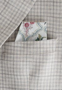 Beige-Anzugjacke aus Stoff mit subtilen Karos, versehen mit einem weißen Taschentuch, das mit floralen Mustern in Pink- und Grüntönen geschmückt ist.