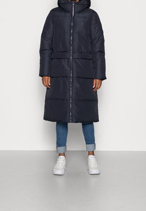 Personne portant un long manteau noir matelassé avec capuche, un jean bleu retroussé et des baskets blanches, debout sur un fond uni.