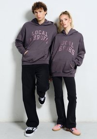 Młody mężczyzna i kobieta w pasujących szarych bluzach z kapturem "Local Heroes" oraz czarnych spodniach, stojący na tle białej ściany.