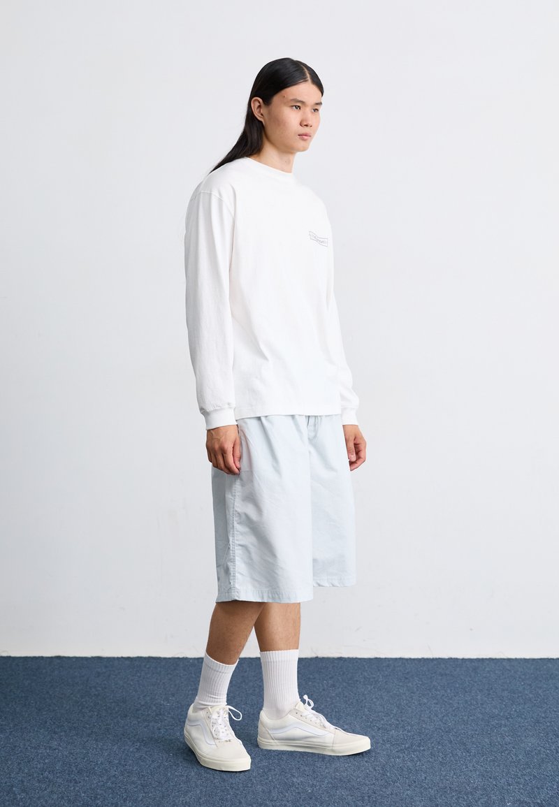 Langärmliges weißes Baumwollhemd mit einem kleinen Grafik, kombiniert mit hellblauen Shorts. Cremefarbene Sneakers und weiße Socken runden das Outfit ab.