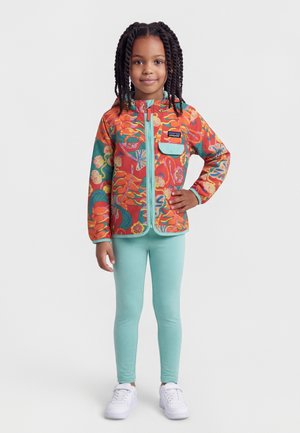 Enfant aux cheveux tressés portant une veste colorée à motif floral, des leggings vert menthe et des baskets blanches, se tenant avec une main sur la hanche.