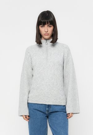 Donna con capelli scuri e lisci che indossa un maglione grigio chiaro a treccia con zip frontale e jeans blu, su uno sfondo bianco.
