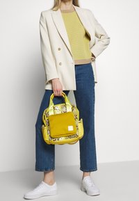 Bolso amarillo con detalles en estampado de serpiente, diseño estructurado, dos asas cortas y un bolsillo frontal. La modelo lleva un blazer crema, un top de punto amarillo y jeans azules.